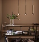 Lampa wisząca Sencillo 3-punktowa w kolorze mosiężnym, z trzema cylindrycznymi kloszami zawieszonymi na czarnych przewodach. Widoczny bukiet roślin w wazonie na stole, na którym znajdują się naczynia i szkło. Tło w ciepłym odcieniu.