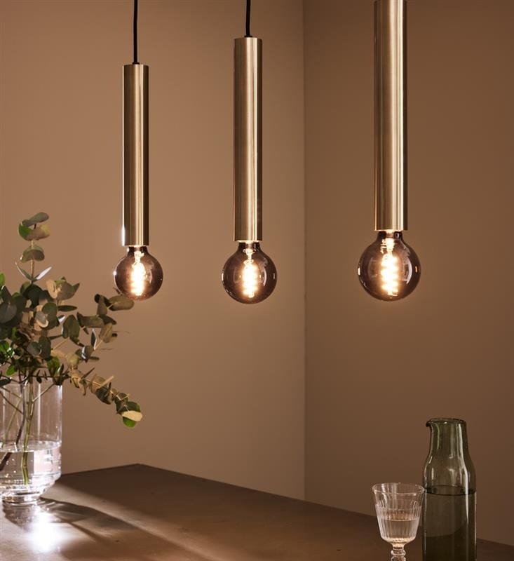 Lampa wisząca Sencillo 3-punktowa w kolorze mosiądzu z trzema długimi, cylindrycznymi oprawami. Każda oprawa posiada żarówkę z przezroczystą kulistą lampą. Produkt zawieszony nad drewnianym blatem, w tle widoczne rośliny i szklane naczynia.