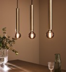 Lampa wisząca Sencillo 3-punktowa w kolorze mosiądzu z trzema długimi, cylindrycznymi oprawami. Każda oprawa posiada żarówkę z przezroczystą kulistą lampą. Produkt zawieszony nad drewnianym blatem, w tle widoczne rośliny i szklane naczynia.