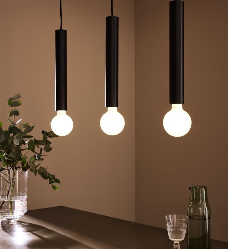 Lampa wisząca Sencillo w kolorze czarnym, z trzema cylindrycznymi oprawami na żarówki, zawieszona w równych odstępach. W tle widoczna szklana waza z roślinami oraz dwa naczynia na stole. Tło w jasnym kolorze.