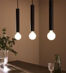 Lampa wisząca Sencillo w kolorze czarnym, z trzema cylindrycznymi oprawami na żarówki, zawieszona w równych odstępach. W tle widoczna szklana waza z roślinami oraz dwa naczynia na stole. Tło w jasnym kolorze.