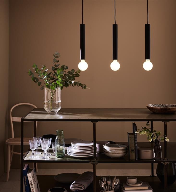 Lampa wisząca Sencillo 3-punktowa w kolorze czarnym, z trzema okrągłymi żarówkami. Zawieszona na cienkich linkach, w nowoczesnym wnętrzu z drewnianym stołem, na którym znajdują się naczynia i dekoracje roślinne. Tło w ciepłym odcieniu.