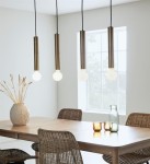 Lampa wisząca Sencillo 4-punktowa w kolorze mosiądzu, z czterema cylindrycznymi oprawkami i okrągłymi żarówkami. Zawieszona nad drewnianym stołem w jasnym wnętrzu z oknem, otoczona naturalnymi dekoracjami.