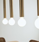 Lampa wisząca Sencillo 4-punktowa w kolorze mosiądzu z czterema okrągłymi, białymi żarówkami. Ujęcie z bliska na detal lampy, zawieszonej na prostych, metalowych rurkach. Tło neutralne, podkreślające nowoczesny design.