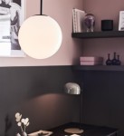 Lampa wisząca Dione 1-punktowa w kolorze czarnym i białym. Okrągła, matowa kula zawieszona na cienkim kablu, umieszczona w nowoczesnym wnętrzu z półkami. Tło w odcieniach różu z dodatkowymi akcesoriami dekoracyjnymi.