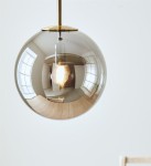 Lampa wisząca Dione w mosiężnym wykończeniu z dymionym, szklanym kloszem w kształcie kuli. Widoczna żarówka wewnątrz, wisząca z sufitu, na białym tle. Klosz odbija otoczenie, co podkreśla jego elegancki design.