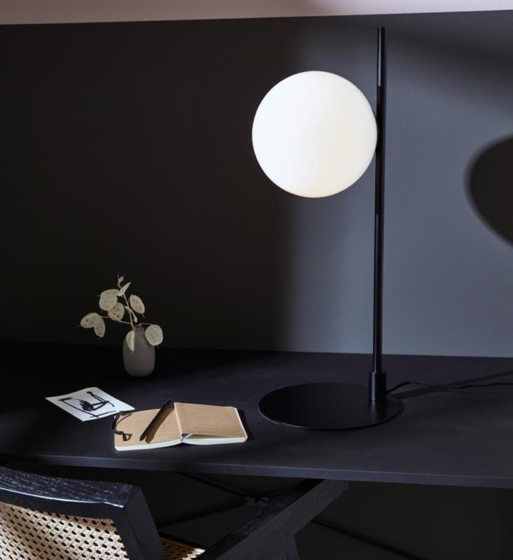 Lampa stołowa Dione 1-punktowa w kolorze czarnym i białym. Okrągła, mleczna kula na smukłym czarnym stelażu, stojąca na okrągłej podstawie. W tle widoczny stół z notatnikiem, długopisem i dekoracyjną rośliną.