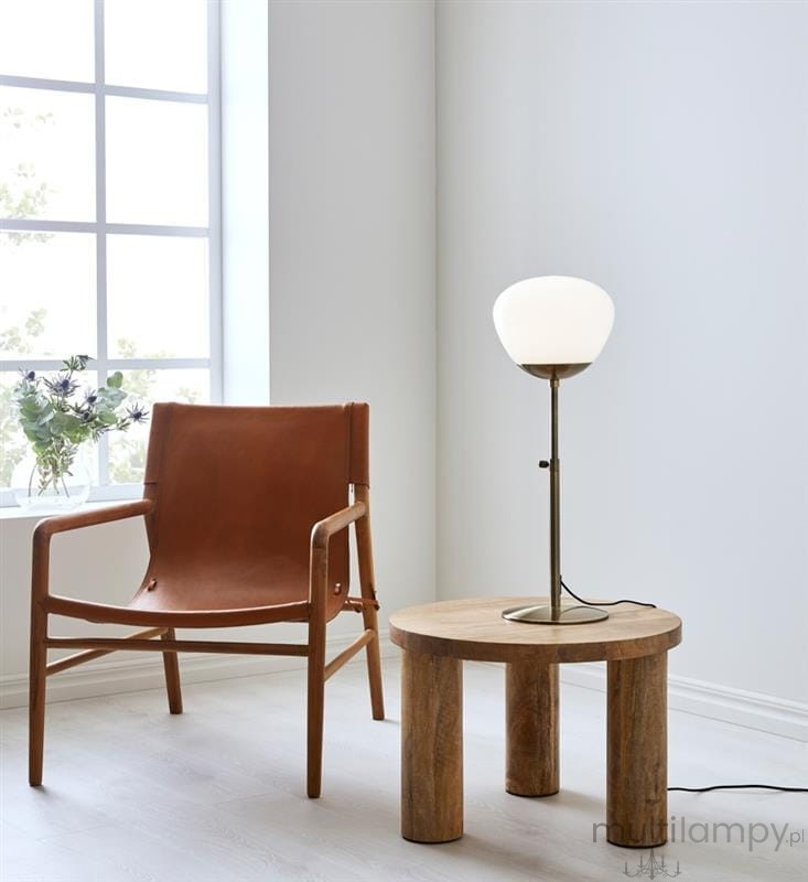 Lampa stołowa Rise 1-punktowa w kolorze antycznego mosiądzu z białym kloszem, ustawiona na okrągłym, drewnianym stoliku obok skórzanego fotela. Tło w jasnym kolorze, okno z widokiem na roślinność. Kabel lampy widoczny na podłodze.