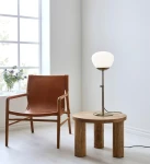 Lampa stołowa Rise 1-punktowa w kolorze antycznego mosiądzu z białym kloszem, ustawiona na okrągłym, drewnianym stoliku obok skórzanego fotela. Tło w jasnym kolorze, okno z widokiem na roślinność. Kabel lampy widoczny na podłodze.