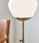 Lampa stołowa Rise 1-punktowa w antycznym mosiądzu z białym kloszem w kształcie kuli. Widok z boku, z widoczną regulacją wysokości i detalami metalowymi. Tło jasne, w tle fragment skórzowego krzesła.