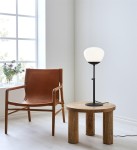 Lampa stołowa Rise 1-punktowa w czarnej podstawie z białym kloszem, ustawiona na okrągłym, drewnianym stoliku. W tle widoczny skórzany fotel i okno z roślinami, na jasnym tle.