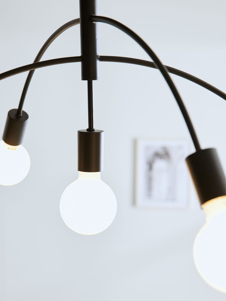Czarna lampa sufitowa 5-punktowa Crux z zaokrąglonymi kloszami, umieszczona na elastycznych ramionach. Widok z dołu, ukazujący trzy źródła światła. Tło jasne, minimalistyczne, z elementami dekoracyjnymi na ścianie.