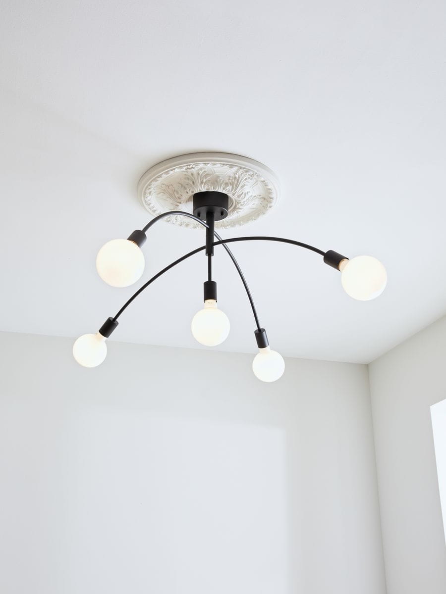 Czarna lampa sufitowa Crux 5-punktowa z okrągłymi kloszami, zamontowana na białym stropie z dekoracyjną rozeta. Wisi w centralnej części pomieszczenia, z pięcioma źródłami światła rozmieszczonymi na elastycznych ramionach.