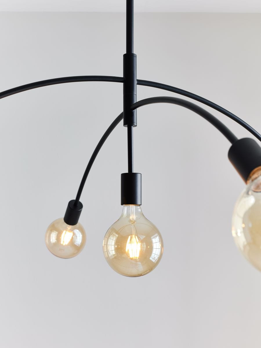 Lampa wisząca Crux pięciopunktowa w kolorze czarnym. Produkt z pięcioma zaokrąglonymi kloszami w stylu retro, zawieszony na czarnych, elastycznych ramionach. Ujęcie z góry, ukazujące detale lampy i jej ciekawe kształty. Tło jasne, jednolite.