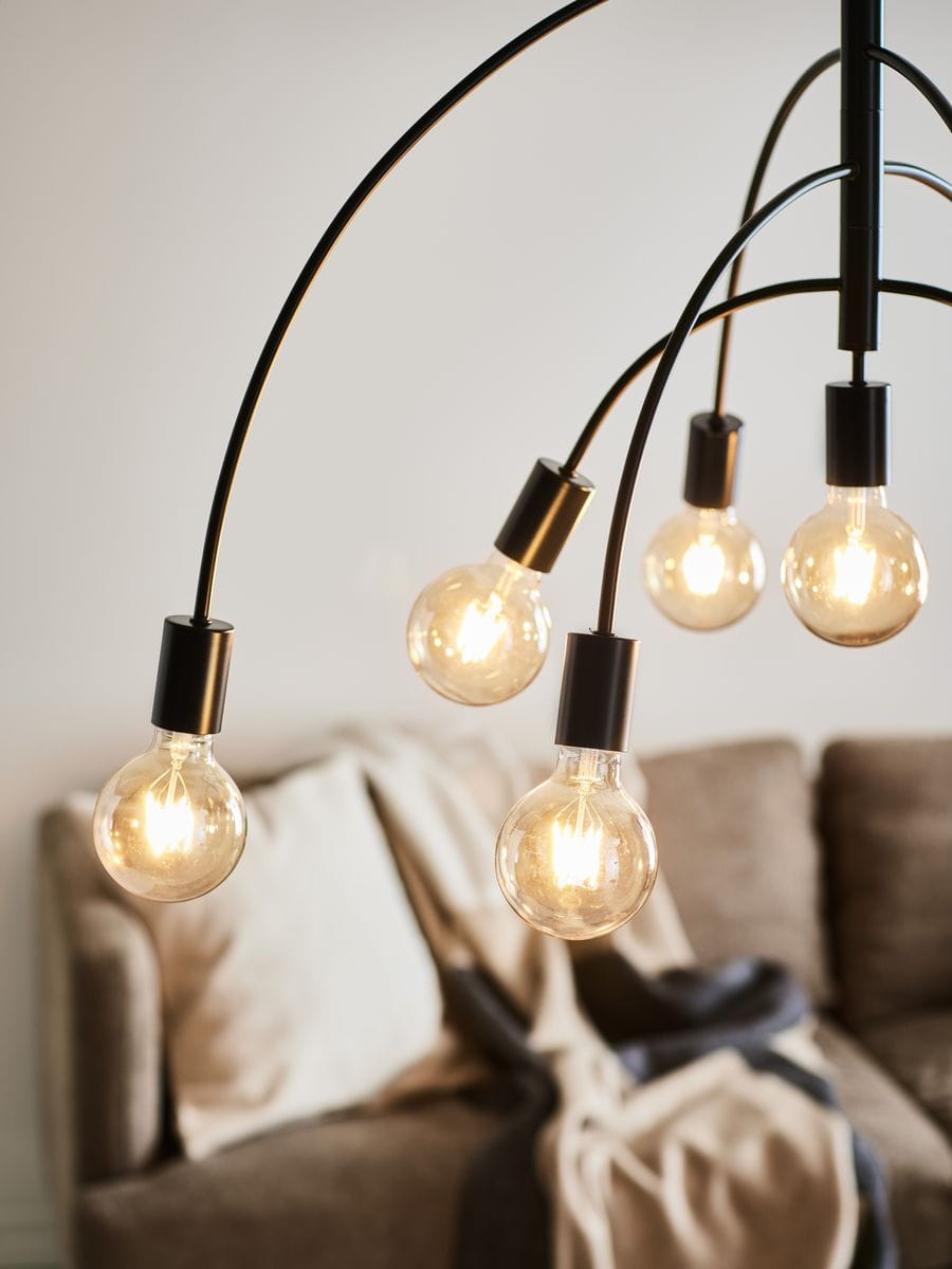 Czarna lampa wisząca Lavello z 9 punktami świetlnymi, z widocznymi żarówkami w przezroczystych kloszach. Ujęcie z góry, z lampą zwisającą nad jasnobrązową kanapą z poduszkami i kocem w tle. Nowoczesny design z delikatnymi łukami.