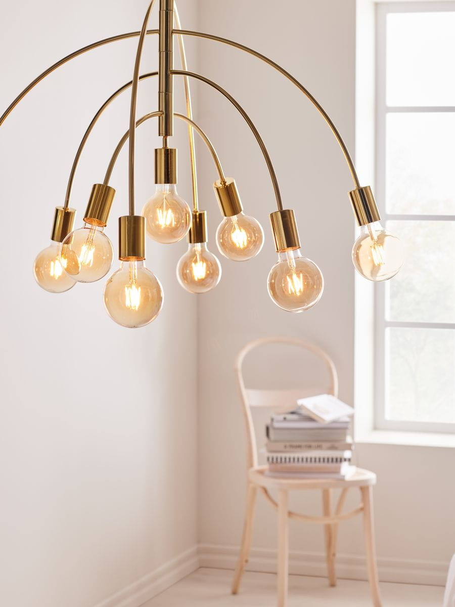 Lampa wisząca Lavello 9-punktowa w kolorze mosiądzu, z siedmioma żarówkami w szklanych kloszach, zawieszona na łukowatych ramionach. Tło jasne, z drewnianym krzesłem i książkami w aranżacji.