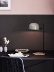Lampa stołowa Pose 1-punktowa w kolorze czarnym i szarym z zaokrąglonym kloszem. Ustawiona na czarnym biurku, obok notatek i dekoracyjnych elementów. W tle jednolite ściany w odcieniach różu i czerni.