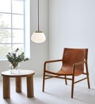 Lampa wisząca Rise 1-punktowa w kolorze antycznego mosiądzu i białym kloszu, zwisająca z ciemnego przewodu. W tle widoczny skórzany fotel i okrągły stół z drewnianymi nogami, w jasnym wnętrzu z dużym oknem.