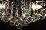 Monte Carlo żyrandol 3-punktowy w kolorze chromu z przezroczystymi, wielokątnymi kryształkami i ozdobnymi łańcuszkami. Widok z bliska, ukazujący detale kryształów oraz źródła światła, na ciemnym tle.