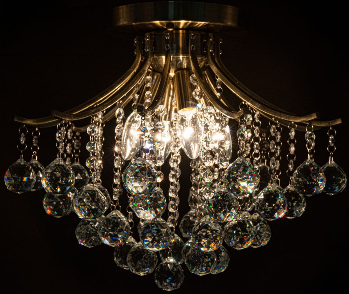 Lampa sufitowa Wenecja 4-punktowa w kolorze mosiądzu, z kryształowymi zwisami. Wyposażona w cztery źródła światła, z ozdobnymi, szklanymi elementami i łańcuszkami. Ujęcie z dołu, zbliżenie na detale i refleksy światła w kryształach.