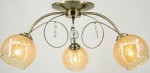 Lampa sufitowa Carmen 3-punktowa w kolorze mosiądzu. Posiada trzy szklane klosze w złotym odcieniu, z dekoracyjnymi kryształkami. Produkt zamontowany na okrągłej podstawie, z lekko zakrzywionymi ramionami. Oświetlenie w stylu klasycznym.
