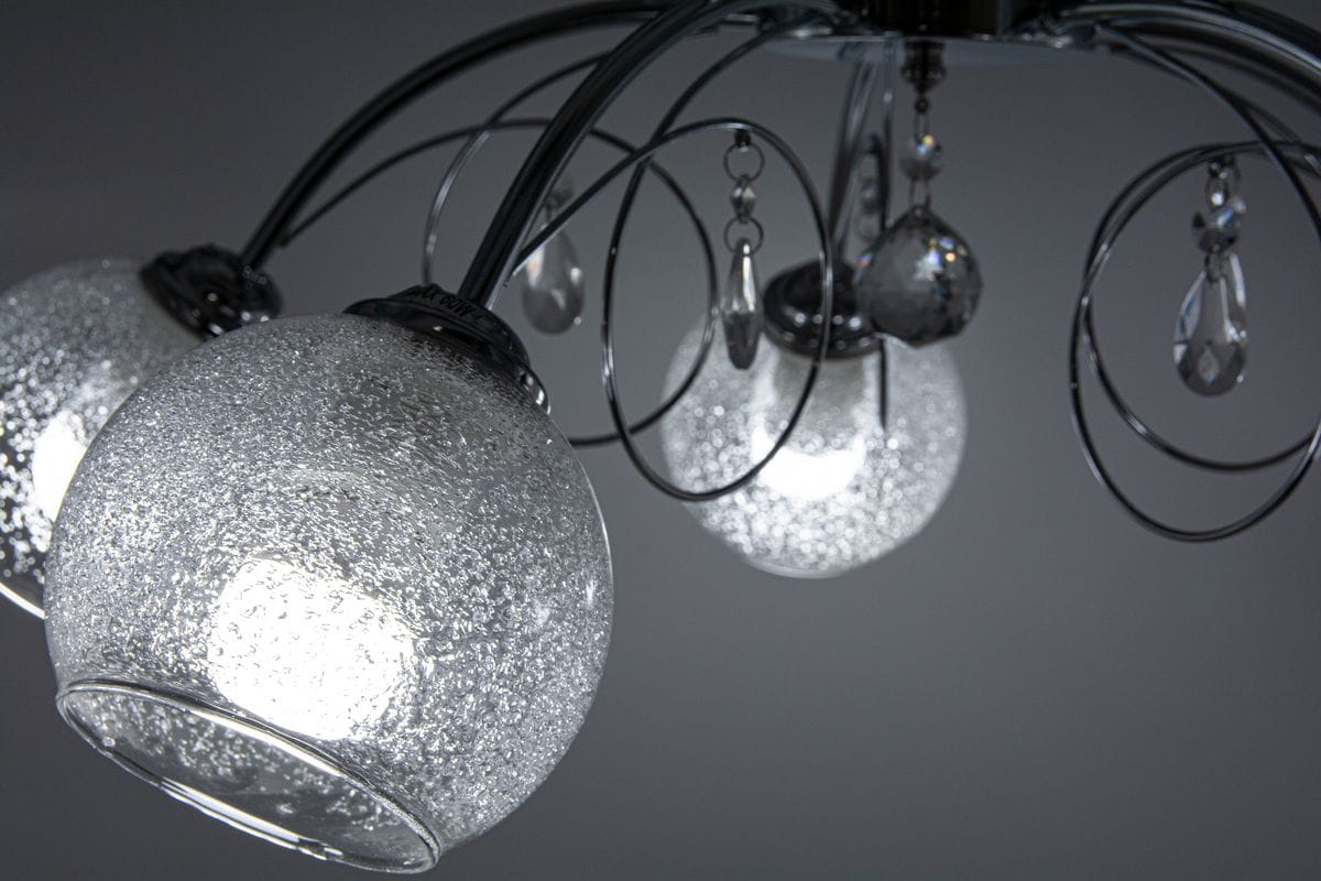 Lampa sufitowa Carmen 5-punktowa w kolorze chromu, posiada pięć kulistych kloszy z przezroczystego szkła z efektem bąbelków. Wisi na stylowych, zakrzywionych ramionach, zdobiona kryształowymi detalami. Ujęcie z bliska na klosze i dekoracje.