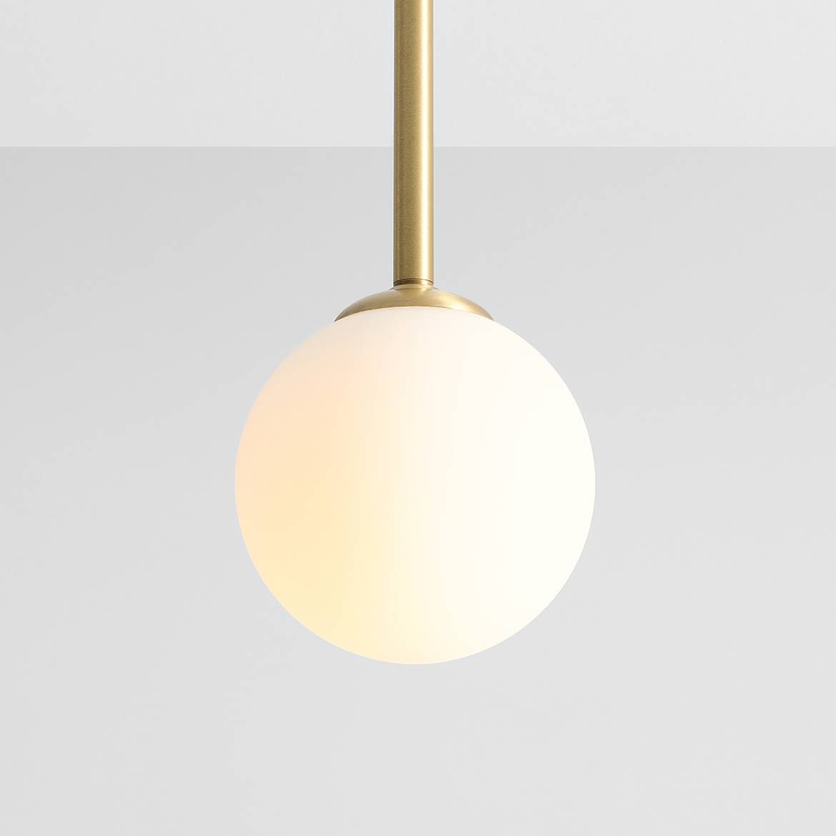 Lampa wisząca Pinne Short w kolorze mosiądzu z jednopunktowym zawieszeniem. Okrągła, matowa kula z białego szkła, zawieszona na cienkim mosiężnym pręcie. Tło jasne, jednolite.