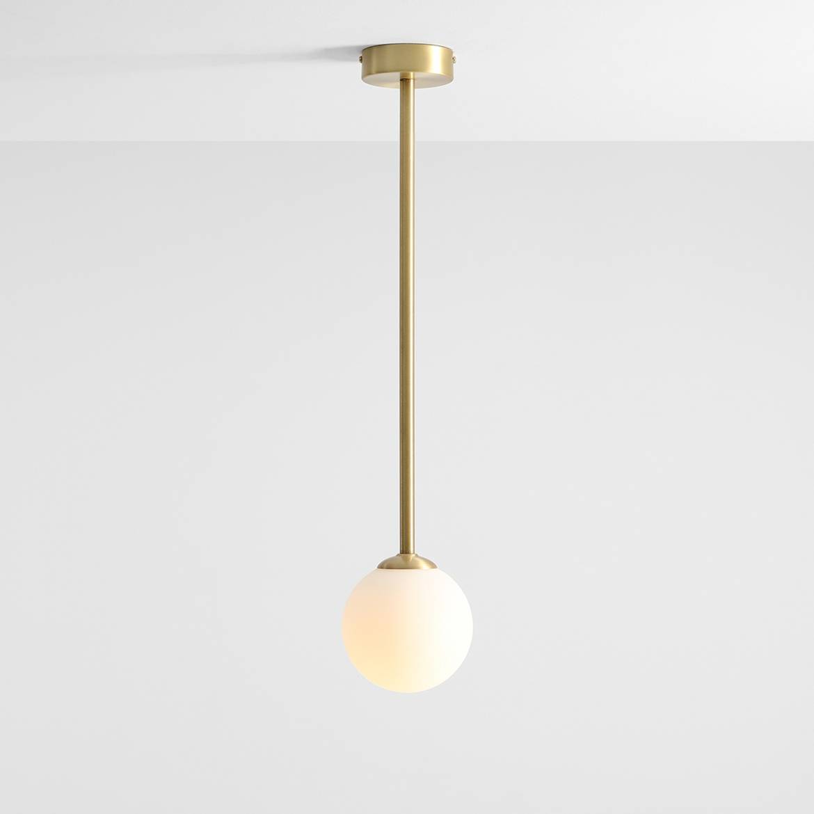 Lampa wisząca Pinne Medium z jednym punktem świetlnym, wykonana z mosiądzu. Posiada długi, prosty metalowy wspornik oraz okrągłą, matową kulę z białego szkła. Zawieszona pod sufitem, na jasnym tle.