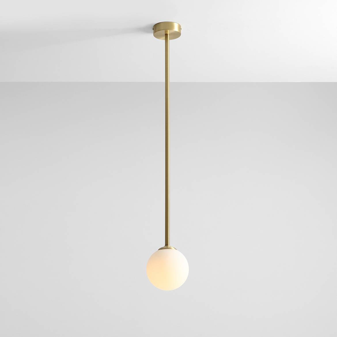 Lampa wisząca Pinne Long w kolorze mosiądzu, z pojedynczym źródłem światła, zawieszona na długim, prostym stelażu. Klosz w formie białej, półprzezroczystej kuli, umieszczony na dolnej części. Tło w jasnym kolorze.