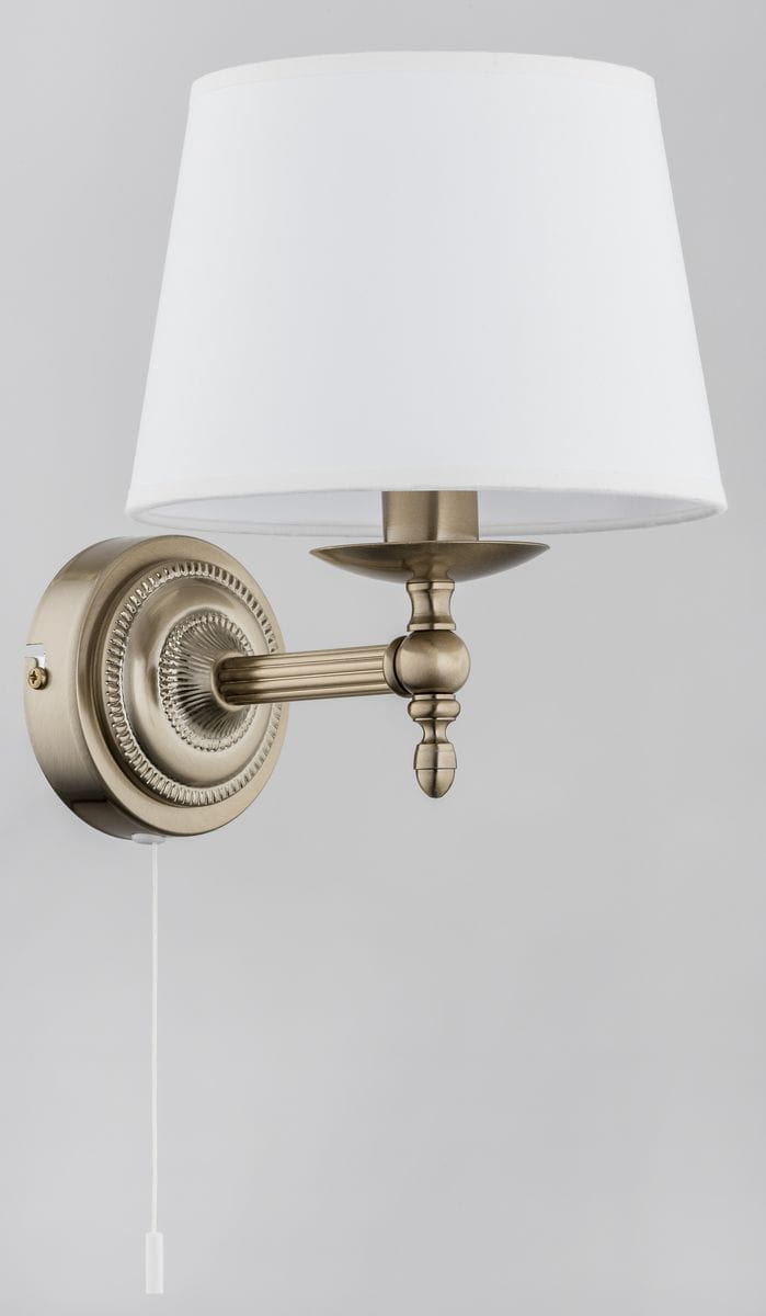 Roksana kinkiet 1-punktowy w kolorze antycznego złota z białym abażurem. Produkt montowany na ścianie, z widocznym detalem w postaci dekoracyjnej podstawy oraz przełącznika. Abażur ma cylindryczny kształt, a całość ma elegancki, klasyczny design.