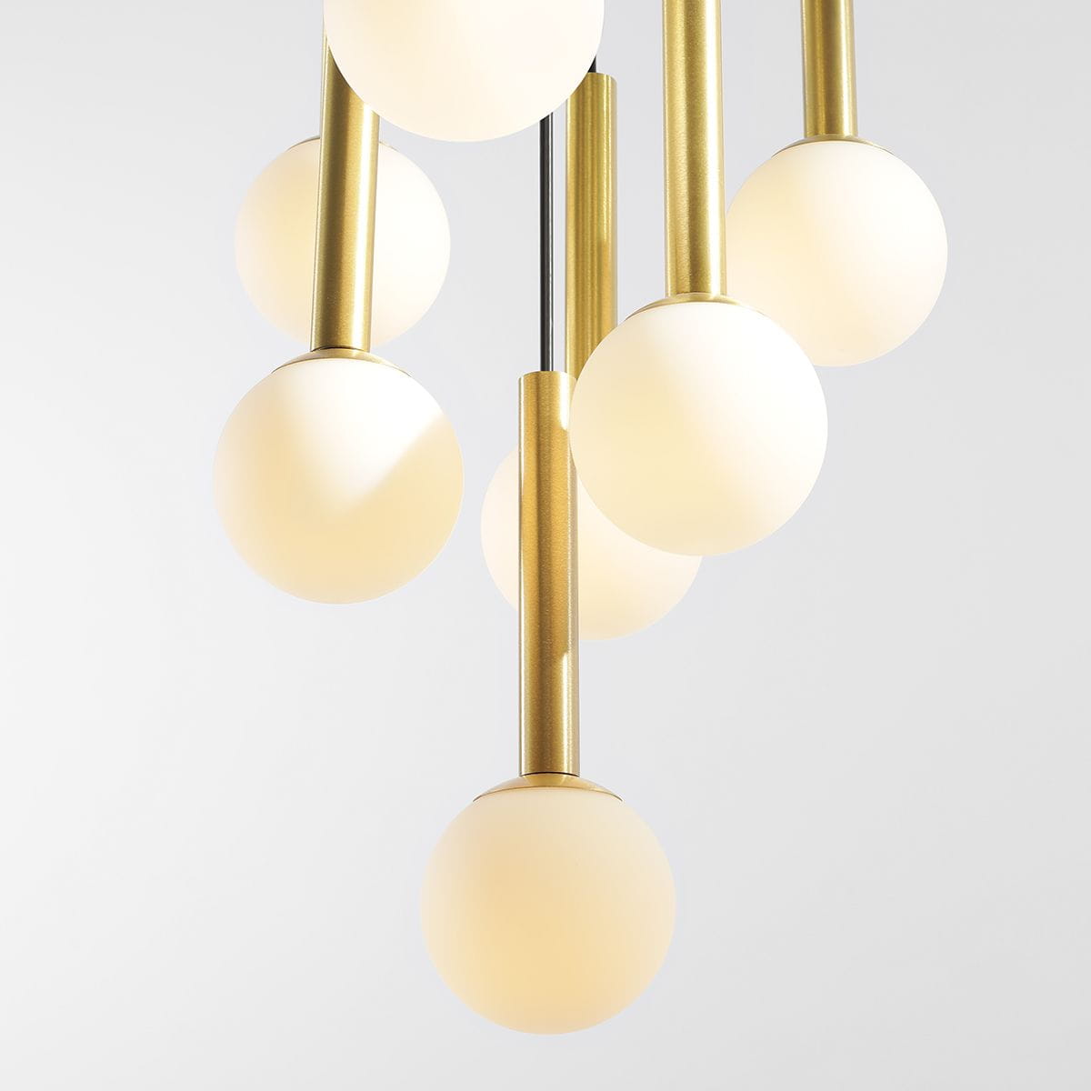 Lampa wisząca Riu 7-punktowa w kolorze czarnym i mosiądzu, z sześcioma okrągłymi, matowymi kloszami w białym kolorze, zamontowana na metalowych prętach w odcieniu mosiądzu, zawieszona w pionie, na jasnym tle.
