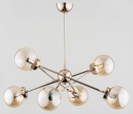 Złota lampa wisząca Sagito z sześcioma kulistymi kloszami, umieszczonymi na metalowych ramionach. Klosze mają przezroczyste, lekko przydymione szkło. Lampa zawieszona na długim wysięgniku, na jednolitym, jasnym tle.