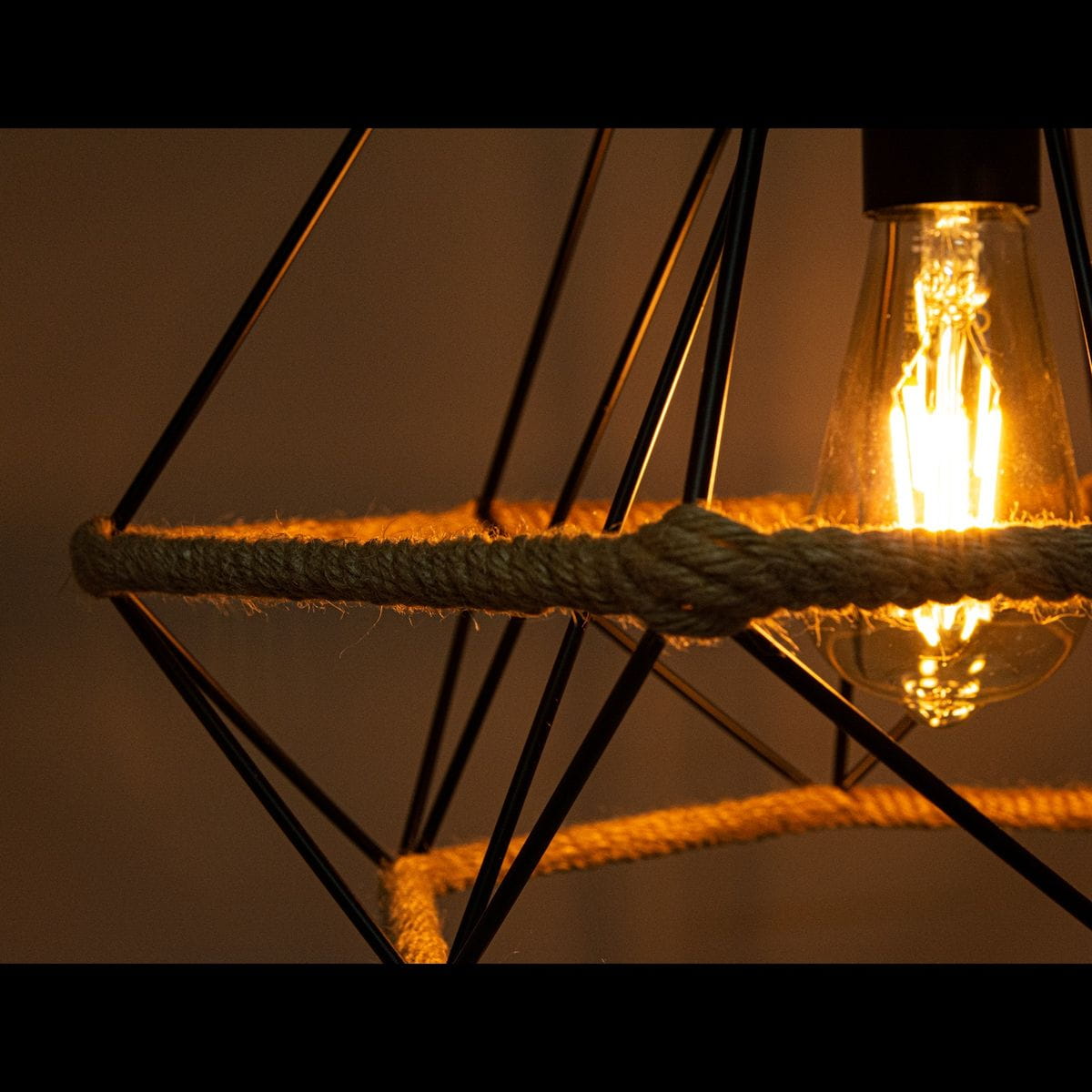 Czarna lampa wisząca Piramida D39 z geometryczną konstrukcją z metalowych prętów. W centralnej części widoczna żarówka otoczona sznurkiem. Lampa ma rustykalny wygląd, idealna do nowoczesnych wnętrz. Zbliżenie na detal i teksturę materiałów.