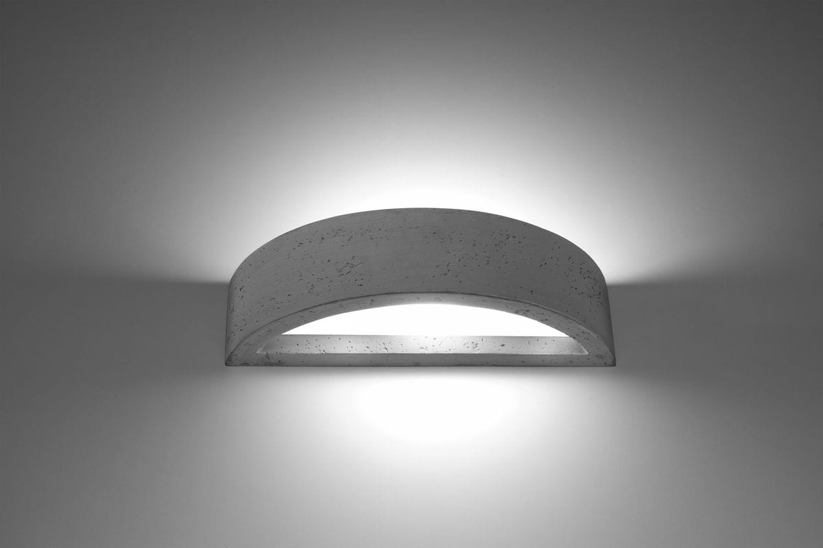 Kinkiet Atena 1-punktowy wykonany z betonu, w kształcie lekko zaokrąglonego prostokąta. Zainstalowany na ścianie, emituje delikatne światło. Powierzchnia ma szaro-beżową fakturę z widocznymi drobnymi wtrąceniami. Tło proste, jednolite.