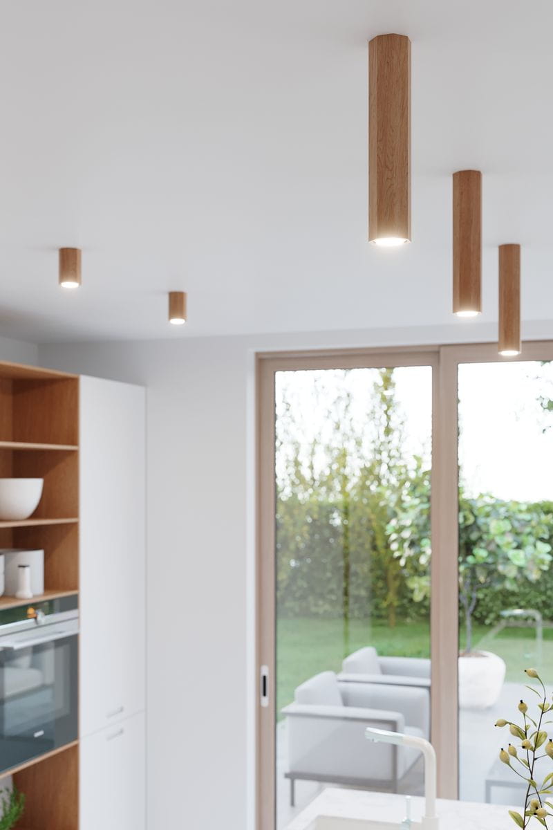 Lampa sufitowa Zeke H30 w kolorze dębowym, w kształcie cylindrów, zawieszona na suficie. Widoczne źródła światła na górze, w nowoczesnym wnętrzu z dużymi oknami. Tło z elementami roślinności i meblami w przestrzeni dziennej.