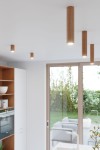 Lampa sufitowa Zeke H30 w kolorze dębowym, w kształcie cylindrów, zawieszona na suficie. Widoczne źródła światła na górze, w nowoczesnym wnętrzu z dużymi oknami. Tło z elementami roślinności i meblami w przestrzeni dziennej.