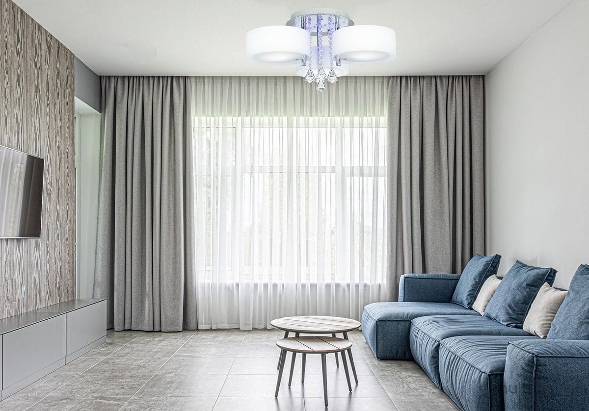 Lampa sufitowa Antila E27 3-punktowa w kolorze chromu i białym, z pilotem. Posiada okrągłe klosze i dekoracyjne elementy z kryształków. Zawieszona w nowoczesnym wnętrzu z szarymi zasłonami i niebieską sofą, na jasnym tle.