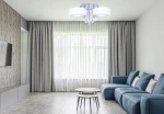 Lampa sufitowa Antila E27 3-punktowa w kolorze chromu i białym, z pilotem. Posiada okrągłe klosze i dekoracyjne elementy z kryształków. Zawieszona w nowoczesnym wnętrzu z szarymi zasłonami i niebieską sofą, na jasnym tle.