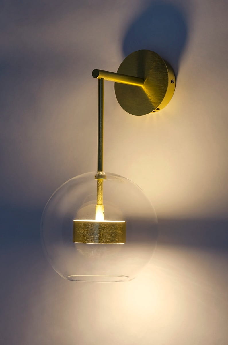 Kinkiet Capri LED 1-punktowy w kolorze złotym, z okrągłym, metalowym mocowaniem. Szklana kula z przezroczystego szkła osłania źródło światła, zawieszona na cienkim, złotym ramieniu. Oświetlenie emituje ciepłe światło na tle ściany.
