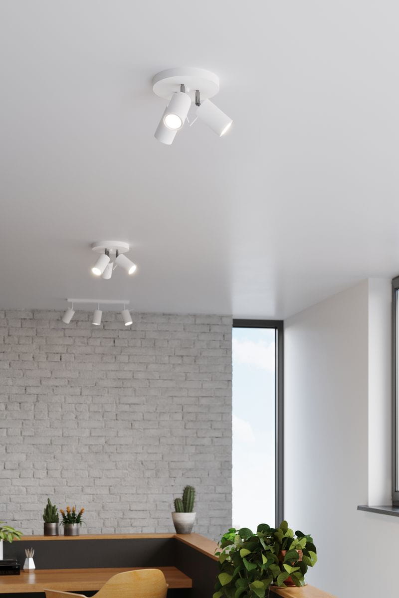 Trójpunktowa lampa sufitowa w białym kolorze, z trzema regulowanymi reflektorami. Ujęcie z góry, zamontowana na jasnym suficie. W tle widoczna ceglana ściana oraz rośliny w doniczkach, tworzące nowoczesny kontekst aranżacyjny.