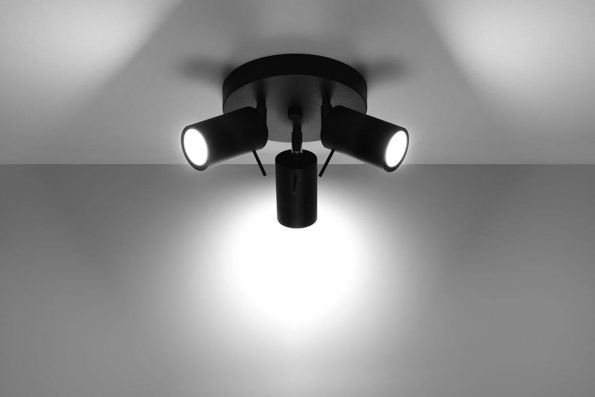 Czarna lampa sufitowa (spot) 3-punktowa z cylindrycznymi reflektorami, zamontowana na okrągłej podstawie. Reflektory skierowane są w różnych kierunkach, emitując intensywne światło. Ujęcie z dołu, na jasnym tle.