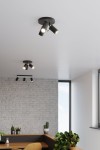 Trzypunktowa lampa sufitowa w kolorze czarnym, zamontowana na suficie, z ruchomymi reflektorami skierowanymi w różne kierunki. W tle widoczne okno i ceglana ściana, co tworzy nowoczesne wnętrze.