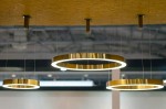 Lampa wisząca Ring D80 LED w złotym kolorze, z okrągłym kształtem i podświetleniem LED. Zawieszona na cienkich linkach, prezentuje nowoczesny design. Widoczne są trzy lampy w różnych wysokościach, umieszczone na drewnianym suficie.