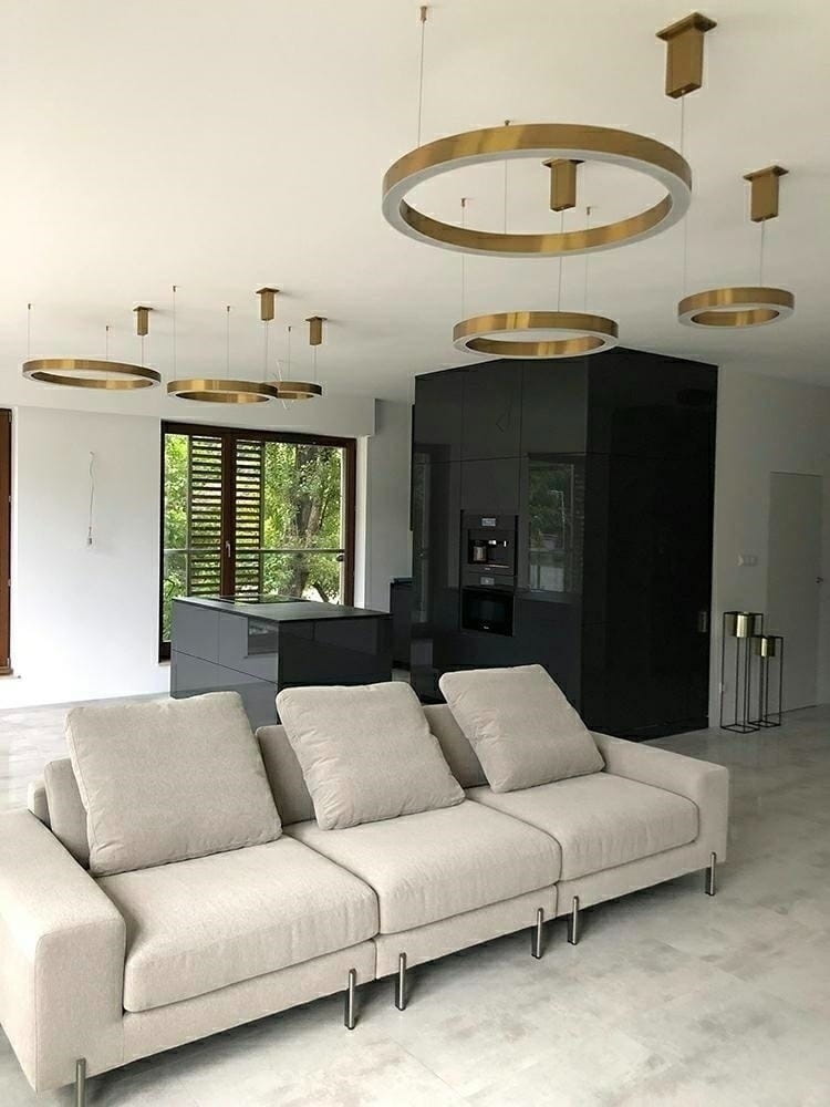 Złota lampa wisząca Ring D80 LED, o okrągłym kształcie, zawieszona nad jasną sofą w nowoczesnym wnętrzu. Widoczne są także inne lampy w tle, a całość znajduje się w przestronnym pomieszczeniu z oknami.