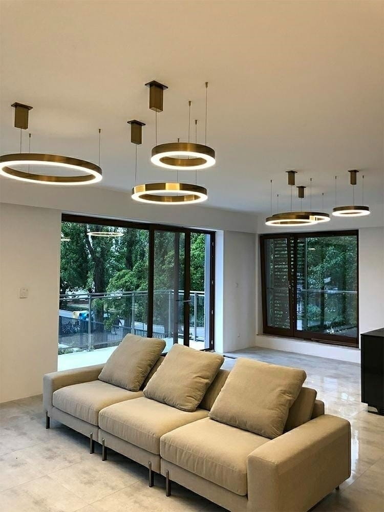 Lampa wisząca Ring D40 LED w kolorze złotym, 1-punktowa, z okrągłym kloszem emitującym białe światło. Zawieszona na cienkich linkach, w nowoczesnym wnętrzu z szarymi płytkami i jasną sofą. Widoczne okna z widokiem na zieleń.