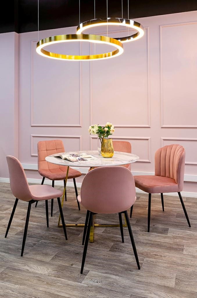 Lampa wisząca Ring D100 LED w złotym kolorze, składająca się z jednego okrągłego pierścienia z diodami LED. Zawieszona nad okrągłym stołem, w jasnym, nowoczesnym wnętrzu z różowymi krzesłami. Tło w odcieniach różu, ściana z dekoracyjnymi panelami.