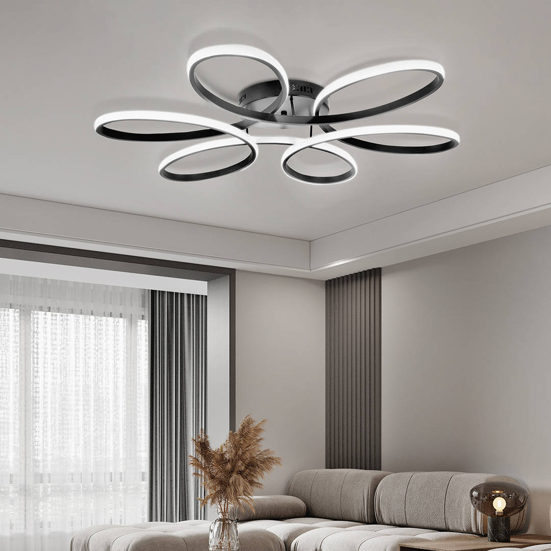 Czarna lampa sufitowa LED 100W w kształcie ośmiu okręgów, z białym podświetleniem. Zamontowana na suficie w nowoczesnym wnętrzu. Elementy dekoracyjne w tle, jasne zasłony i kanapa w kolorze jasnoszarym. Widoczny pilot do zdalnego sterowania.