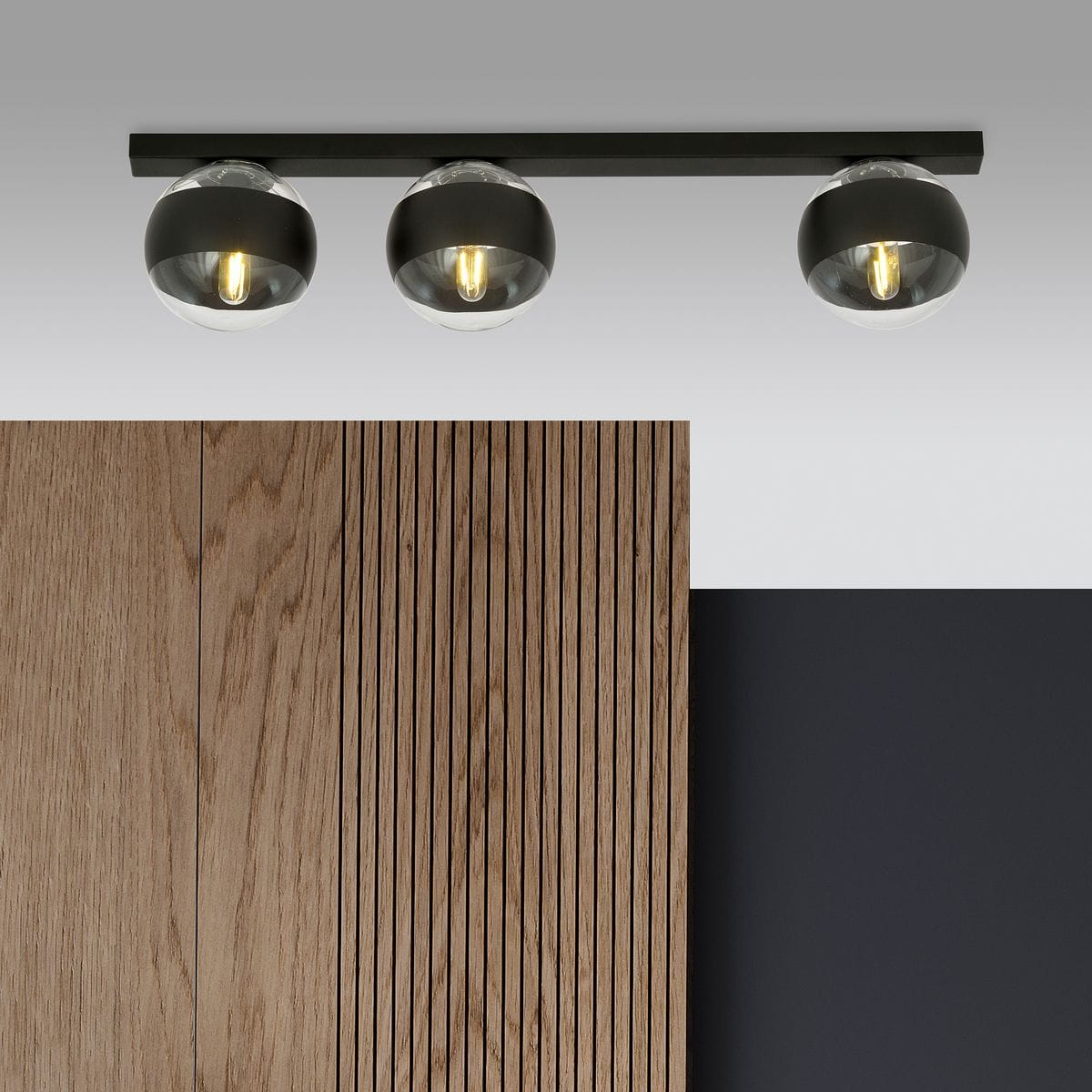 Lampa sufitowa Fit w kolorze czarnym z trzema okrągłymi kloszami, zamontowana na poziomej, prostokątnej listwie. Klosze są przezroczyste z czarnymi akcentami, emitujące ciepłe światło. Tło z drewnianymi panelami i ciemną ścianą.
