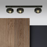 Lampa sufitowa Fit w kolorze czarnym z trzema okrągłymi kloszami, zamontowana na poziomej, prostokątnej listwie. Klosze są przezroczyste z czarnymi akcentami, emitujące ciepłe światło. Tło z drewnianymi panelami i ciemną ścianą.