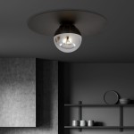 Czarna i grafitowa sufitowa lampa 1-punktowa z okrągłym kloszem, umieszczona na ciemnym tle. Klosz jest szklany, z widocznym źródłem światła wewnątrz. Produkt zamontowany na suficie, w minimalistycznym wnętrzu z szarymi ścianami i prostymi półkami.